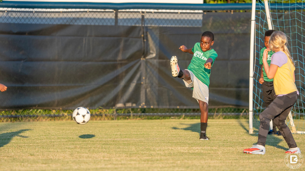 A player in a green DD4 Soccer shirt kicks the ball high as a teammate and an opposing player watch near the goal.