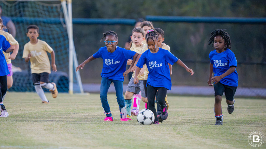 Players in blue DD4 Soccer shirts and gold shirts race toward the ball, with a goal in the background.