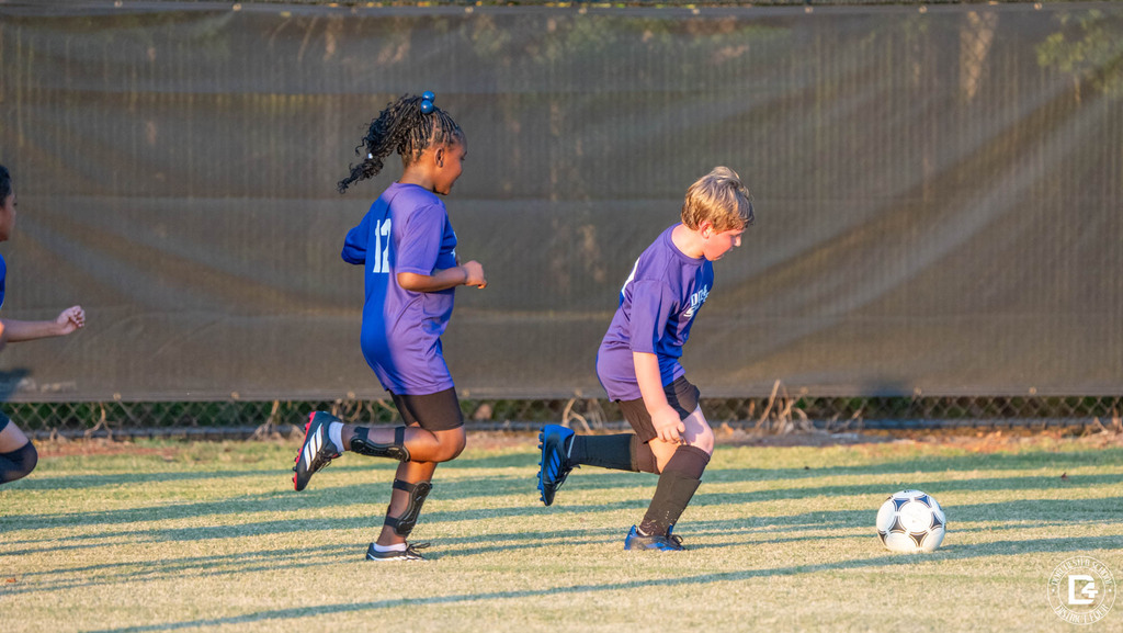 Two players in purple DD4 Soccer shirts run downfield as one dribbles the ball across the grass.