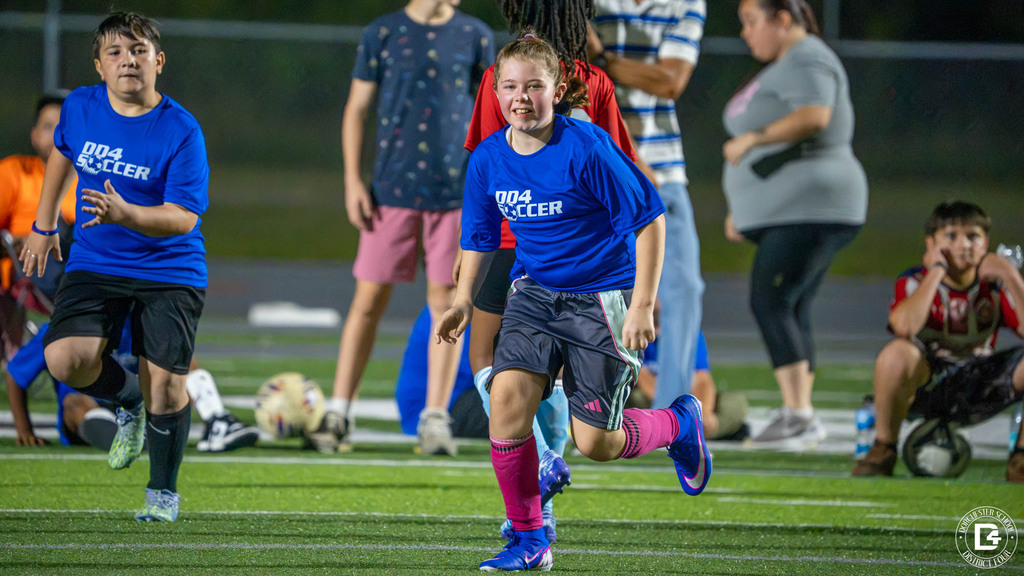 A smiling player in a blue DD4 Soccer shirt runs across the turf field as a teammate follows behind and families watch from the sideline.