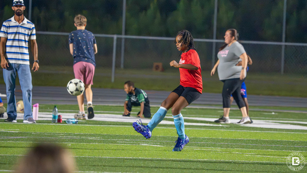 A player in a red DD4 Soccer shirt and light blue socks passes the ball on a turf field during an evening game.