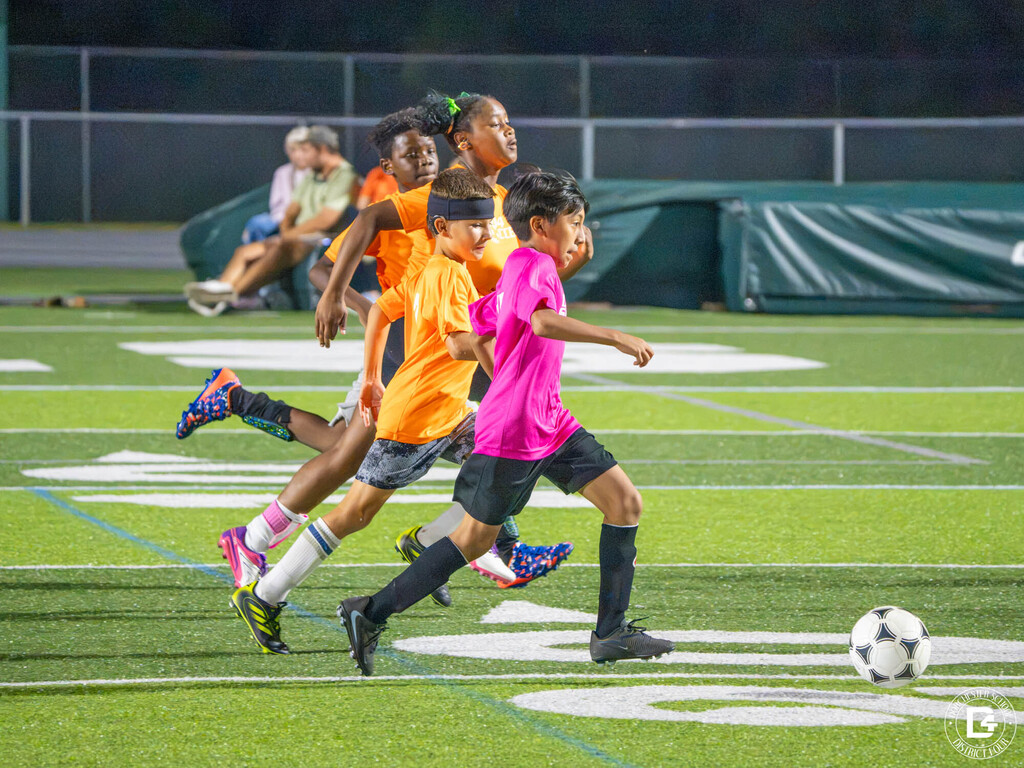 Players in orange and pink shirts sprint after the ball on a lighted turf field during a night game.
