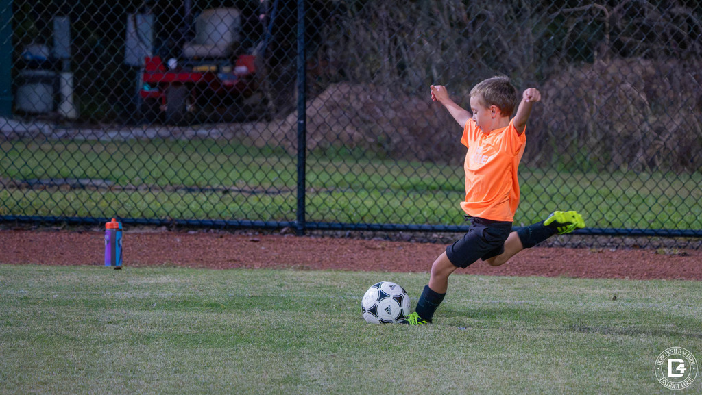 A young player in an orange shirt winds up with arms raised to kick a soccer ball on a grass field.