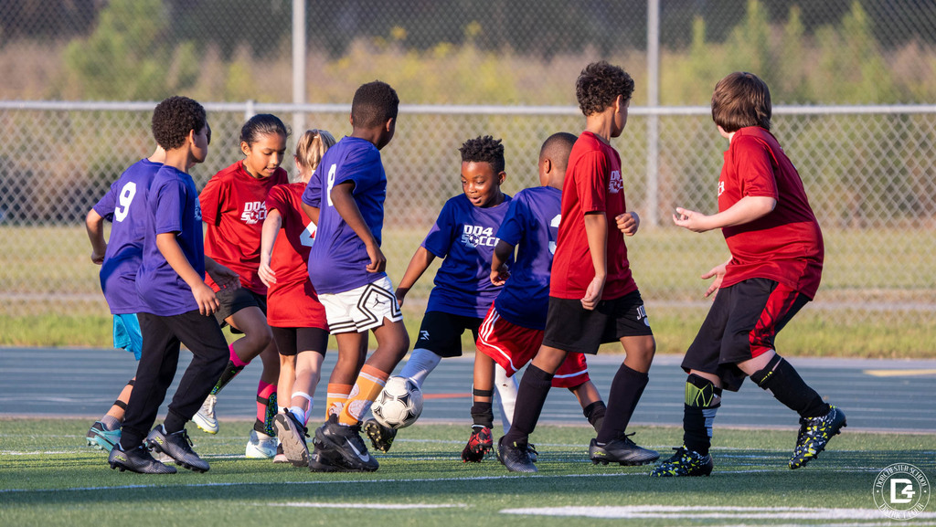 Young soccer players in red and purple DD4 Soccer shirts crowd around the ball during a game on a turf field.