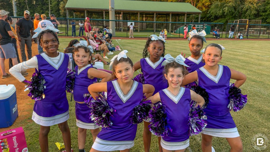 Seven young cheerleaders in purple and white uniforms with silver bows pose with pom-poms on the sideline as families watch the game.