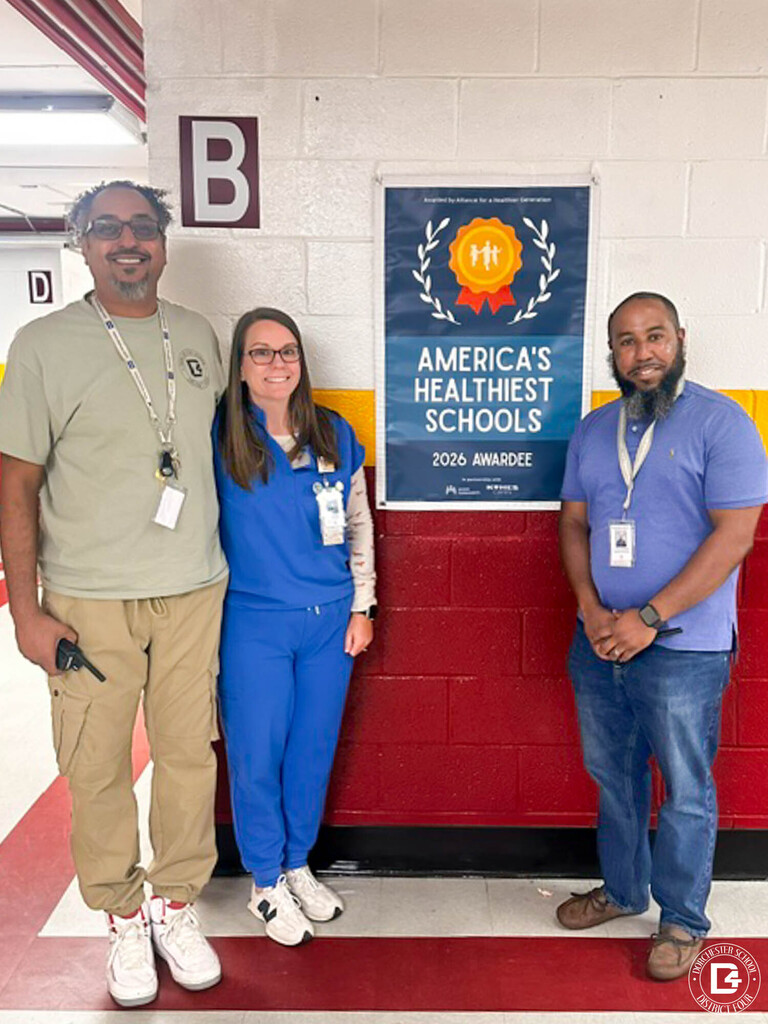 Three smiling Williams Memorial Elementary staff members stand in a school hallway beside a framed America's Healthiest Schools 2026 Awardee poster on a white and red block wall. On the left, a man in a sage green t-shirt and khaki pants stands next to a woman in royal blue scrubs. On the right, a man in a blue polo and jeans stands with his hands clasped. The Dorchester School District Four logo appears in the bottom corner.  If the woman in scrubs is Nurse Peters, you could swap in "Nurse Peters, in royal blue scrubs," and name the other two staff members as well, since names make alt text more meaningful for screen reader users.