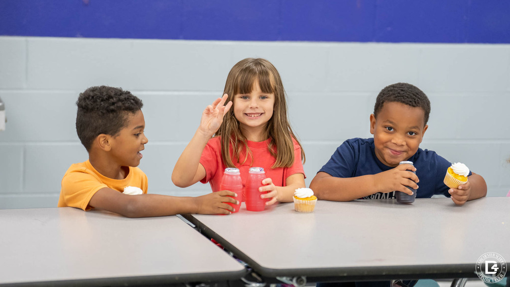 Three young students sit at a cafeteria table with cupcakes and juice. The girl in the middle smiles and waves at the camera.