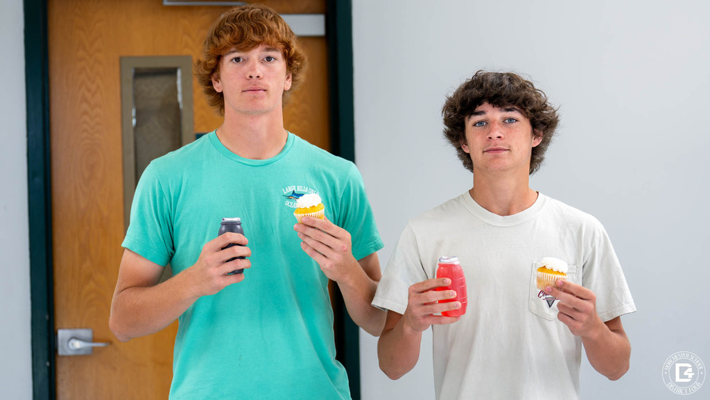 Two high school students stand side by side in a hallway, each holding a cupcake and a juice bottle.