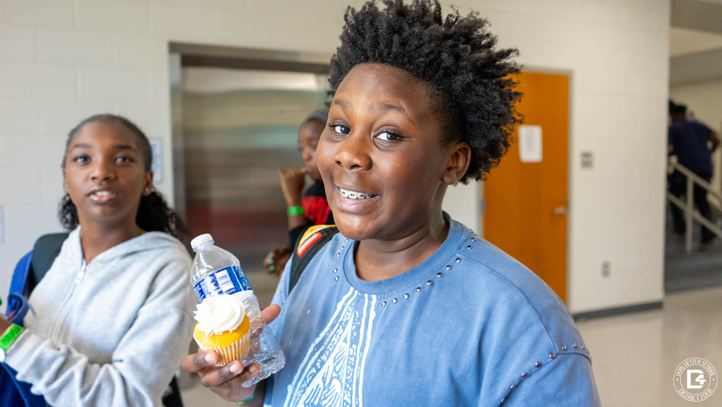 A smiling student holds a white-frosted cupcake and a water bottle in a school hallway as classmates walk by.