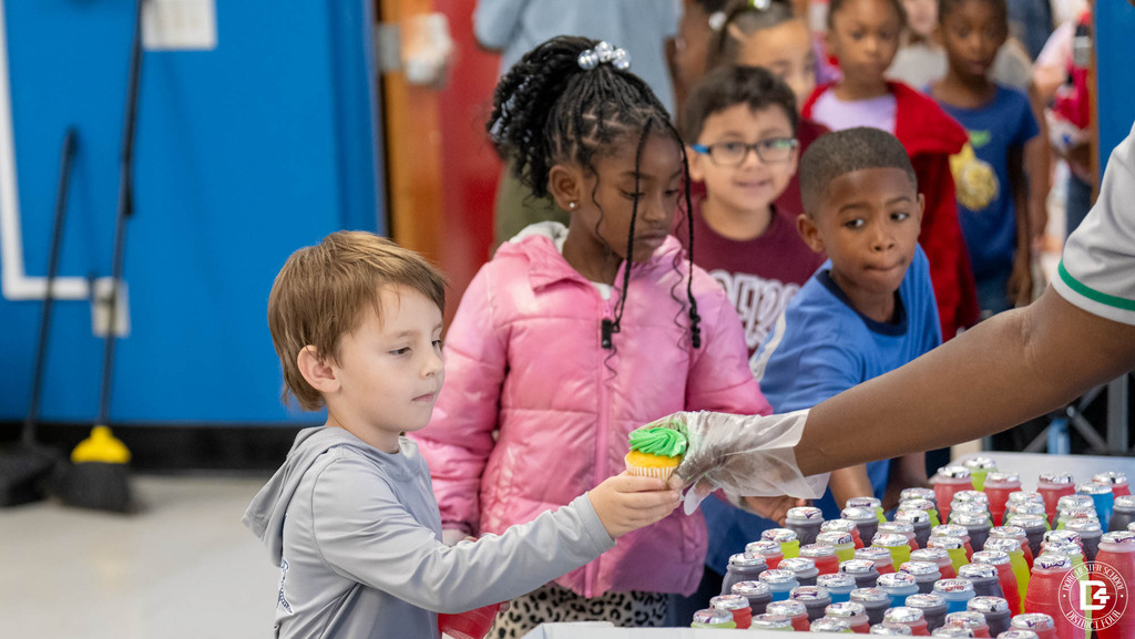 A young student reaches for a green-frosted cupcake handed to him by a staff member, while classmates wait in line behind him next to a table full of colorful juice bottles.