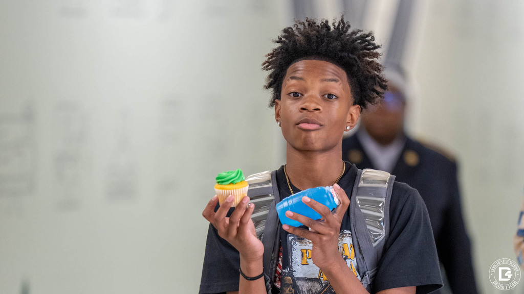 A middle school student with a backpack holds up a green-frosted cupcake and a blue juice bottle, giving the camera a serious look.