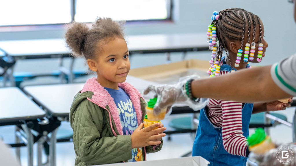 A young student in a pink-lined jacket holds a green-frosted cupcake as a staff member hands out more cupcakes to her classmates.