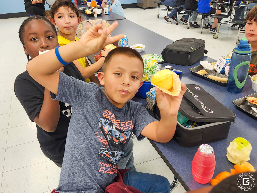 A student in a racing car t-shirt holds up a cupcake and makes a peace sign while sitting at a cafeteria table with classmates.