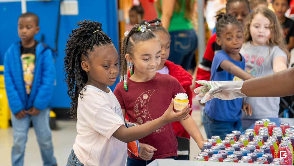 A young student carefully accepts a white-frosted cupcake as smiling classmates line up beside a table of colorful juice bottles.