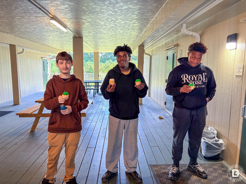Three students stand on a covered wooden walkway outside their classrooms, each holding green-frosted cupcakes.