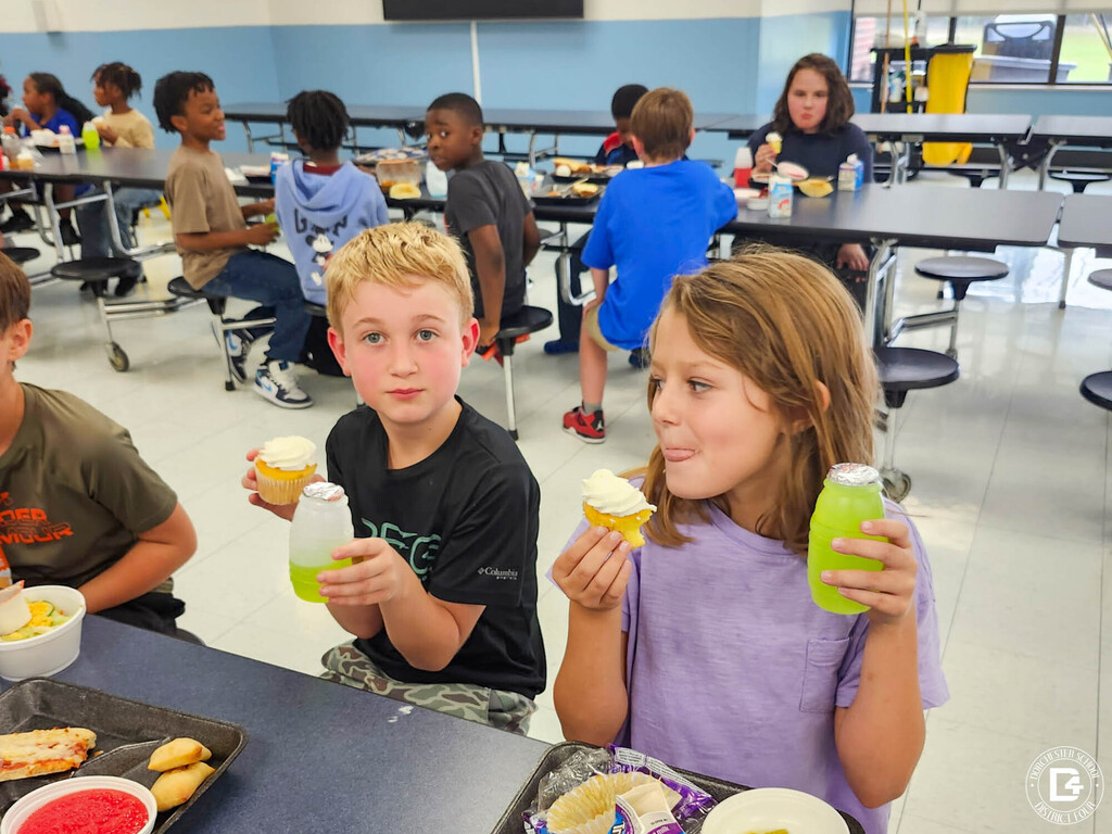Two students sit at a cafeteria table holding frosted cupcakes and green juice bottles. One stares at the camera while the other licks her lips.