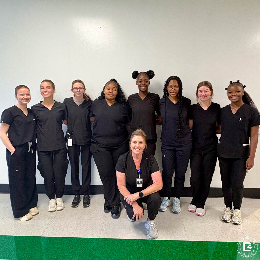 Eight Woodland High School Nursing Assistant students in black scrubs stand in a row against a white wall, smiling. Their instructor, Nurse Reeves, kneels in front of the group in matching black scrubs and a name badge.