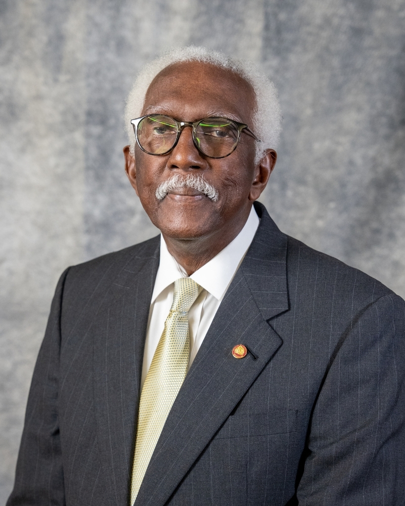 Portrait of Dr. James L. Hodges, Dorchester School District Four Board Member and Board Secretary, smiling softly in a dark pinstripe suit, white shirt, and gold patterned tie with a pin on his lapel, against a gray textured backdrop. The Dorchester School District Four logo appears in the bottom right corner.