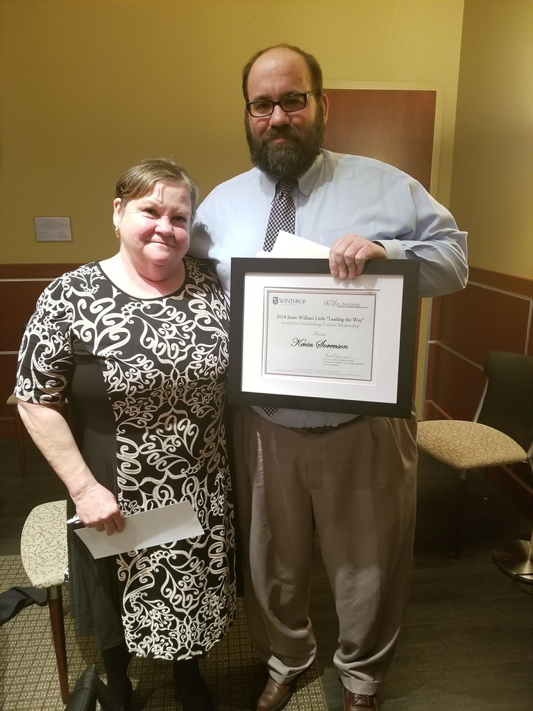 Former DD4 history teacher Kevin Sorenson stands smiling beside a woman in a black and white patterned dress. He wears glasses, a light blue shirt, and a patterned tie, and holds a framed certificate from Winthrop University and The Rex Institute naming him a finalist for the 2018 Jessie William Little “Leading the Way” Award for Outstanding Teacher Mentorship.