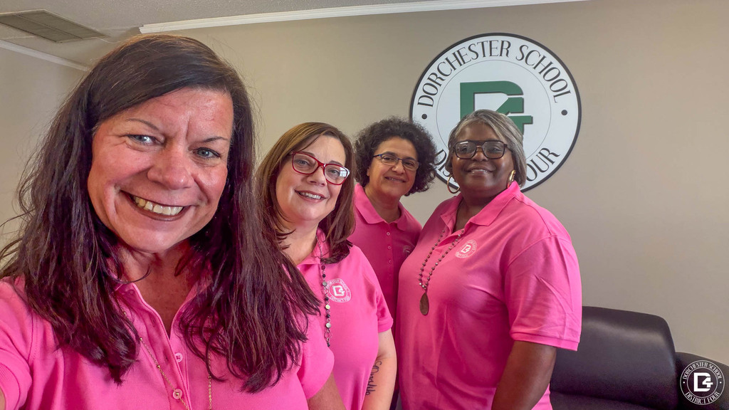 Four smiling staff members in pink DD4 polo shirts take a group selfie in front of the Dorchester School District Four logo on an office wall.