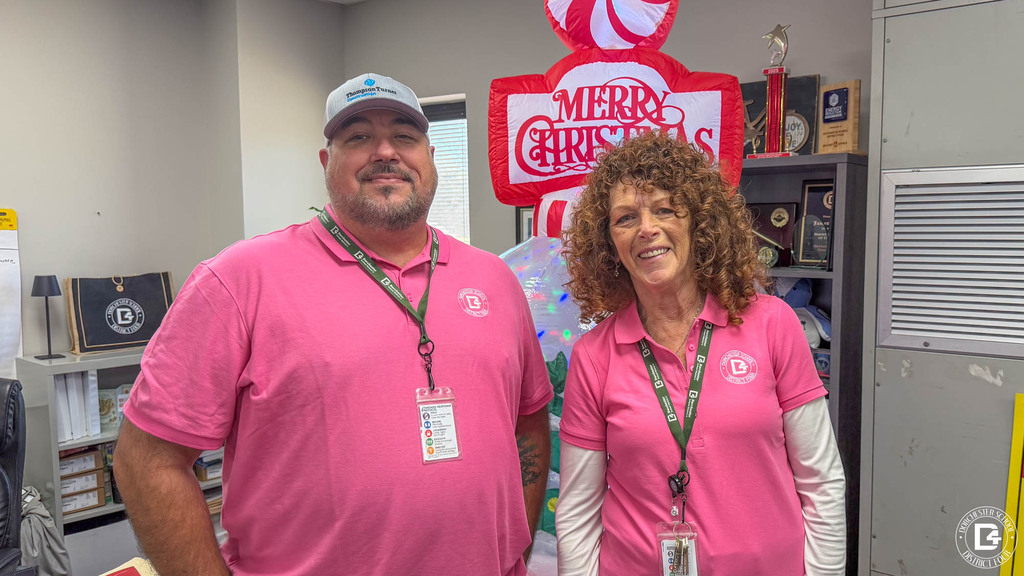 Two staff members in pink DD4 polo shirts and lanyards stand together in an office decorated with an inflatable holiday decoration, trophies, and awards.