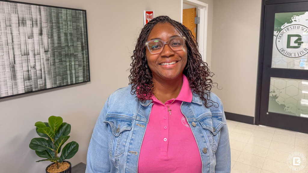 A smiling staff member wearing a pink DD4 polo shirt under a denim jacket stands in the District Office lobby near the Dorchester School District Four logo.