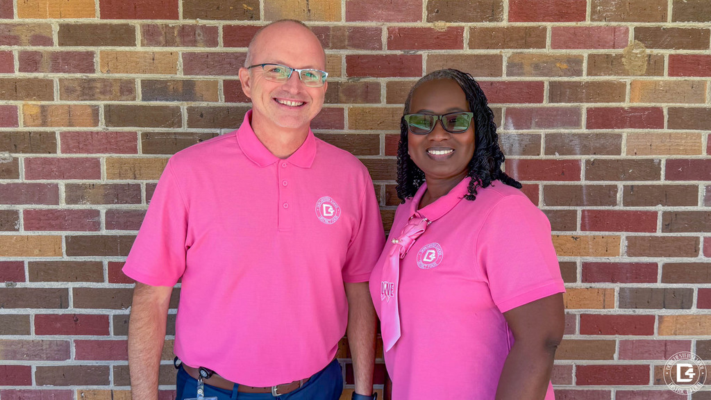 Two smiling staff members in pink DD4 polo shirts pose in front of a brick wall. One wears a pink scarf with a breast cancer awareness ribbon and the word “Love.”