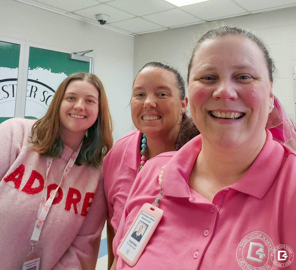 Three smiling District Office staff members take a selfie together in a hallway, two wearing pink DD4 polo shirts and one in a light pink sweater. A Dorchester School District Four logo is visible on a green door behind them.
