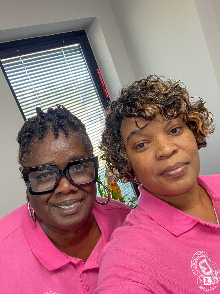 Two staff members in matching pink DD4 polo shirts pose together for a close-up selfie in an office with a window behind them.