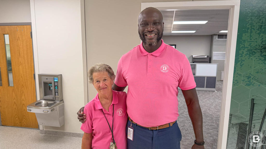 Two staff members in pink DD4 polo shirts pose together in a District Office hallway, one with an arm around the other’s shoulder.