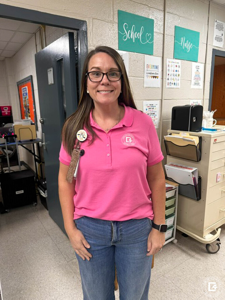 A smiling staff member in a pink DD4 polo shirt and jeans stands in a school nurse’s office, with “School Nurse” signs and a medical cart behind her.