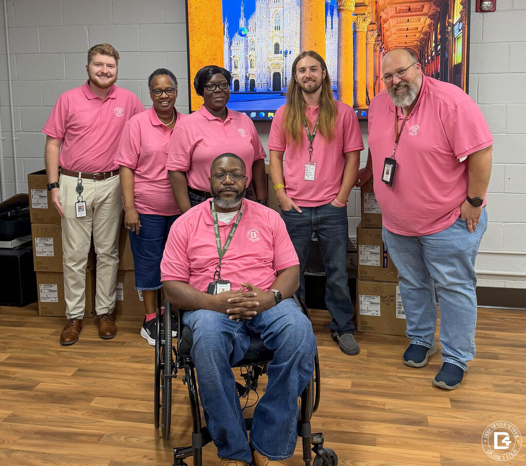 Six District Office staff members in pink DD4 polo shirts pose as a group in front of a large display screen and stacked boxes, with one seated in a wheelchair in front.