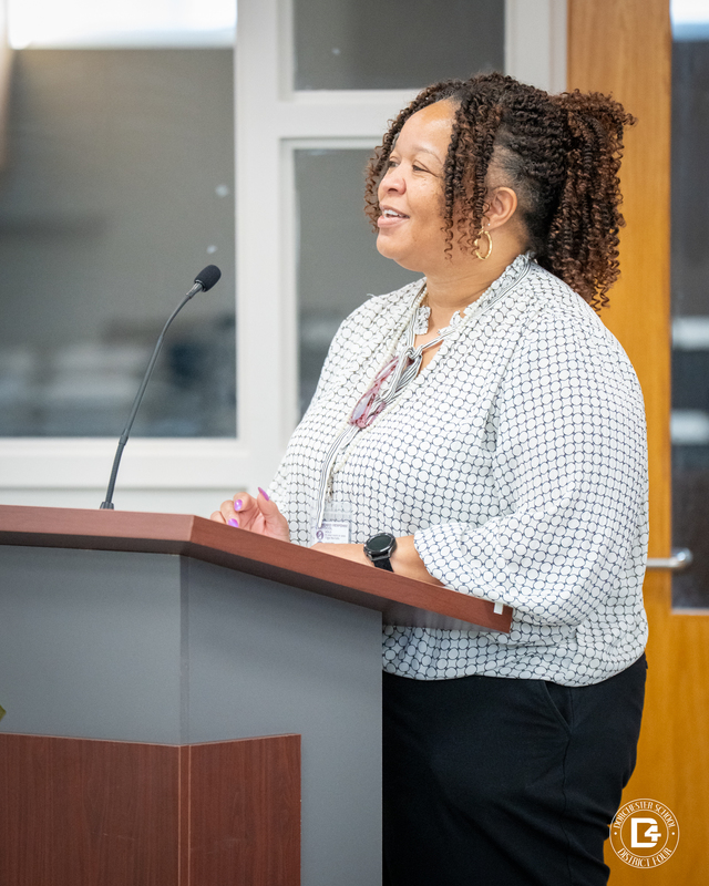 Alisa Lemon, Clay Hill Elementary Principal  woman in a white patterned blouse smiles as she speaks at a podium during the board meeting.