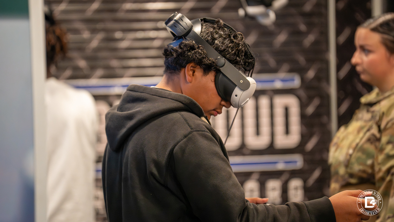 A student wearing a black hoodie uses a virtual reality headset inside the Be Pro Be Proud mobile unit. A blurred student in uniform stands in the background.