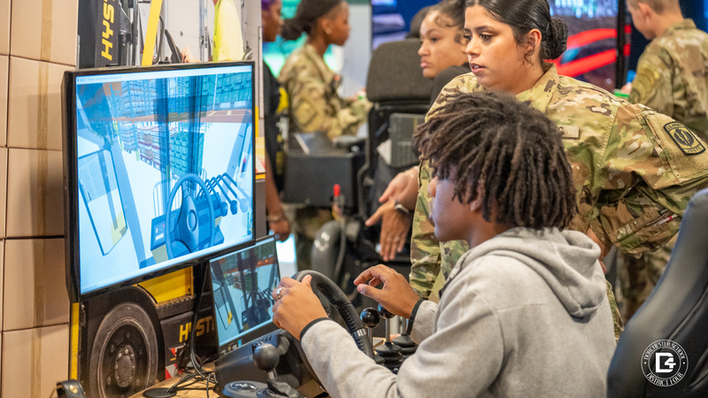 A student sits at a simulator station using a steering wheel and controls to operate a forklift in a virtual warehouse, with a large monitor showing the simulation. A person in military uniform stands nearby offering guidance.