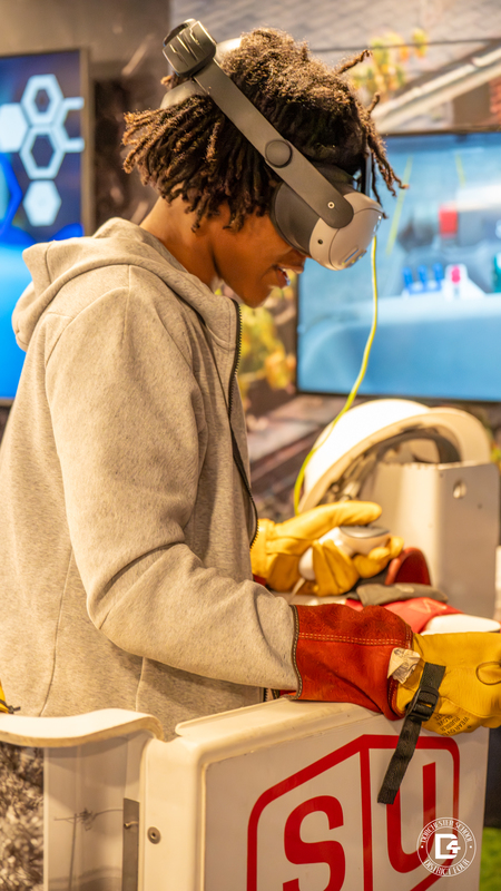 A student wearing a gray hoodie smiles while using a VR headset and protective gloves. He is standing in a mock utility bucket lift, simulating electrical line work.