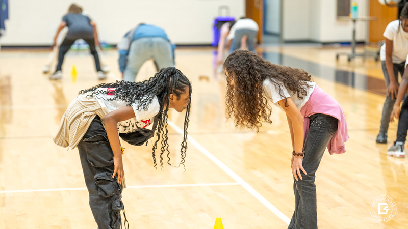 Two girls crouch low and face each other intently during the game, with a yellow cone between them on the gym floor.