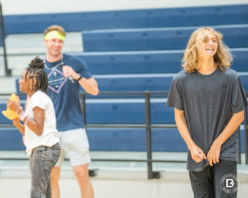 A girl holding an orange cone smiles while standing next to a boy in a gray shirt, with a PE teacher in the background also smiling.