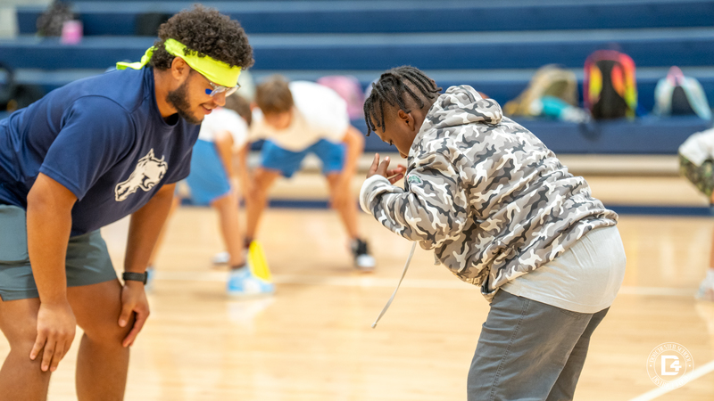 A PE teacher in a blue shirt and headband leans forward as a student in a camouflage hoodie crouches down, both fully engaged in the game.