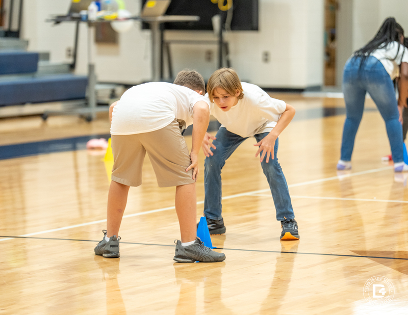 Two boys face each other in a crouched stance, focusing on a blue cone placed on the gym floor between them during the “Head, Shoulders, Knees, and Cones” activity.