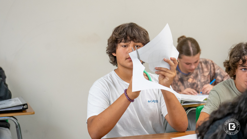 A student in a white shirt carefully cuts out a paper template with scissors as part of a classroom activity.