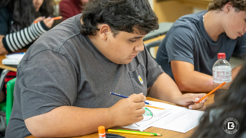 A student in a dark t-shirt colors a green leaf diagram on paper with colored pencils during the lesson.