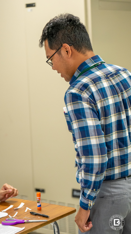 Teacher Mr. Paras, wearing glasses and a plaid shirt, leans over a student’s desk to provide guidance during the activity.