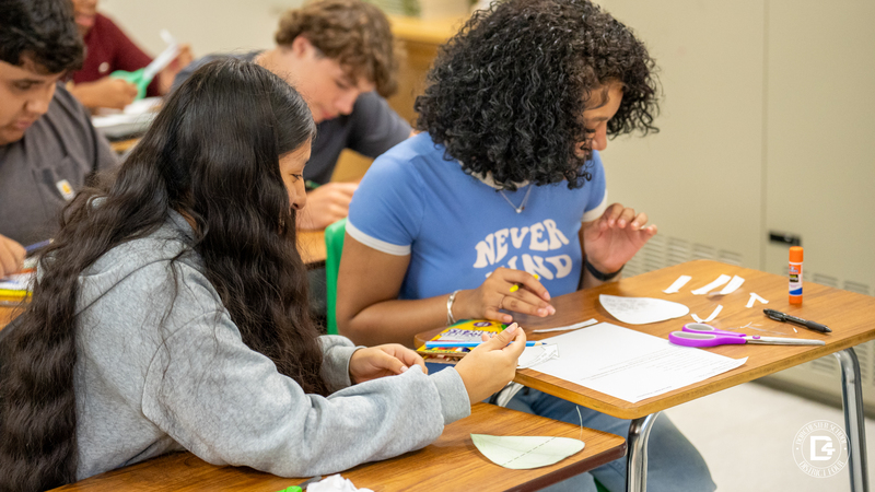 Two students sit side by side at desks, using scissors, glue, and colored pencils to build their photosynthesis leaf models.