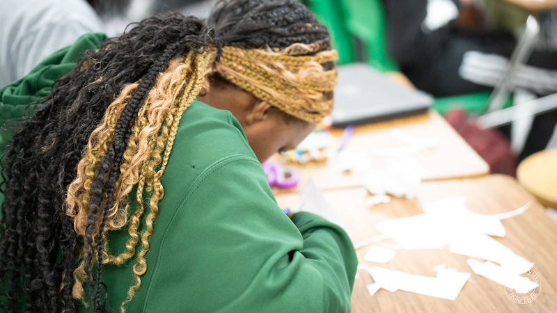 A student in a green hoodie with braided hair works intently at their desk, surrounded by paper scraps from cutting.