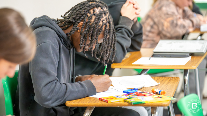 A student in a black hoodie leans over his desk, focused on coloring and labeling his leaf diagram.