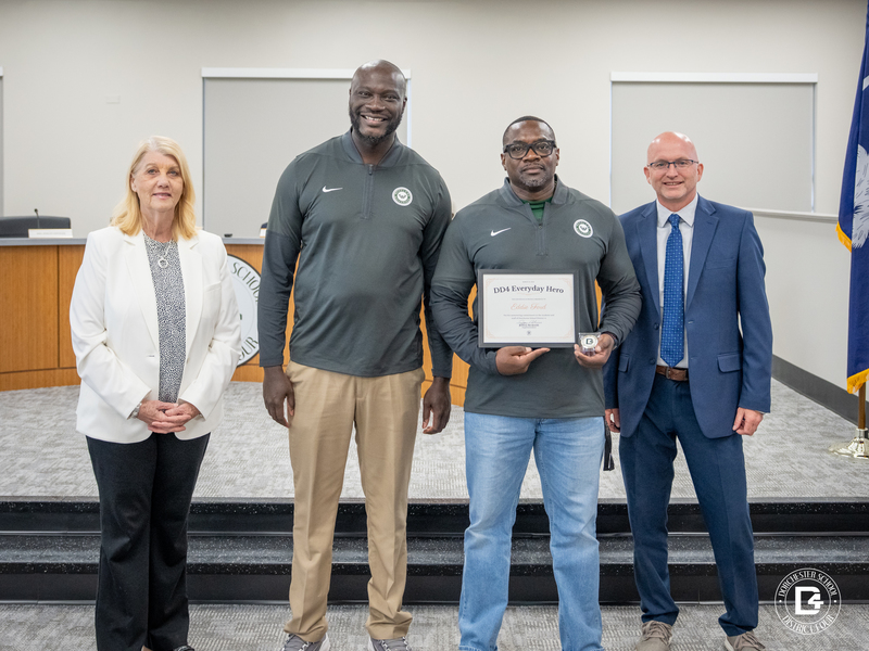 Four adults stand in a school board room as a man holds a framed DD4 Everyday Hero certificate for Eddie Ford along with a small award, with others standing beside him and a Dorchester School District Four logo visible in the corner.