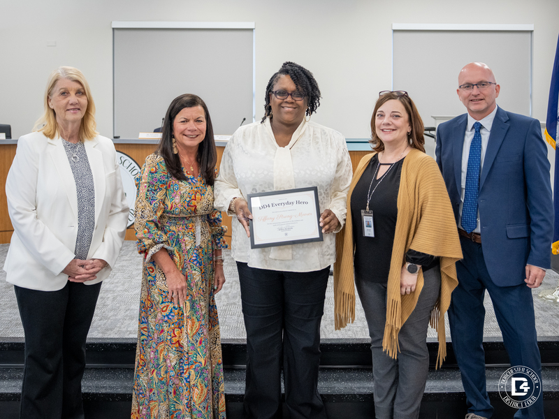 Five adults stand in a school board room as a woman at center holds a framed DD4 Everyday Hero certificate for Tiffany Strong-Moorer, with others standing beside her in support and a Dorchester School District Four logo visible in the corner.
