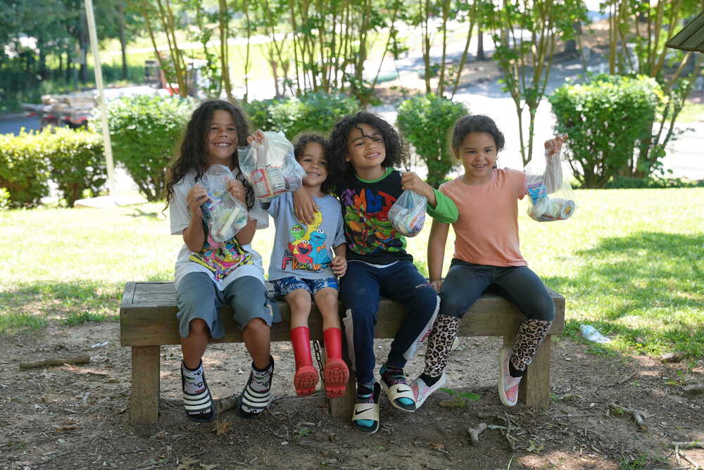 Students hold up their summer meals while sitting on a bench.