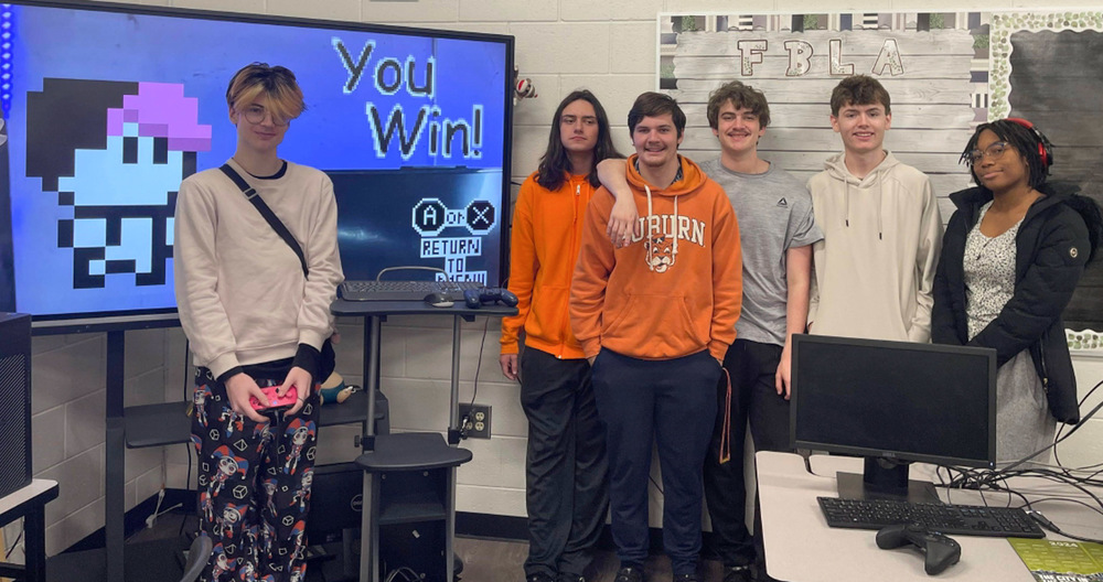Alexander programming students stand in front of a game project. The projects are part of the students' PGAS Cumulative Projects, a culminating experience in which they design and develop original games.